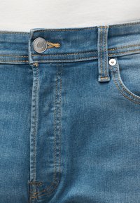 Κοντινό πλάνο στη μέση μπλε τζιν παντελονιού με μεταλλικό κουμπί, θηλιά για ζώνη και ορατές ραφές.