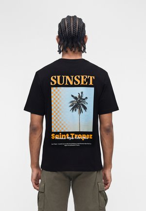 Άτομο με πλεξούδες φοράει μαύρο μπλουζάκι με το κείμενο "SUNSET", γραφικό φοίνικα και λεζάντα "Saint Tropez" στην πλάτη.