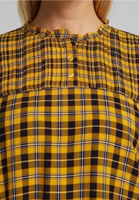 Blusa a cuadros giallo e nera con tessuto testurizzato, scollatura arricciata e cinque bottoni frontali. Il design include motivi a quadro contrastanti.