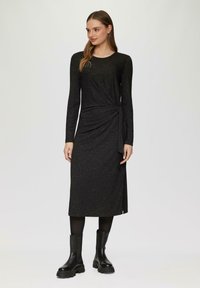 Schwarzes langärmliges Kleid mit einem drapierten Taillendetail, knielang, aus strukturiertem Material mit subtilen Glitzerpartikeln. Kombiniert mit schwarzen Ankle Boots.