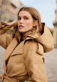 Bežim trench coat s velikom kapuljačom; izrađen od glatke tkanine, ima strukiran pojas i rever ovratnik. Model ima labavu, valovitu kosu.