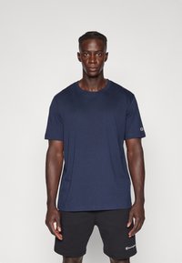 T-shirt en coton bleu marine à col rond et manches courtes, avec un petit logo sur la manche gauche et une texture lisse.