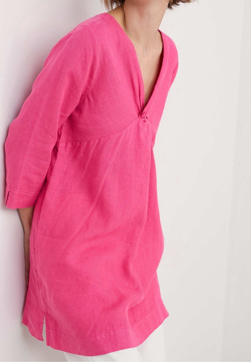 Femme portant une robe rose vif à manches longues avec un col en V et un détail de nœud, appuyée contre un mur blanc, les mains dans le dos.
