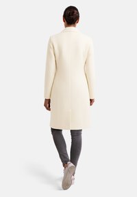 Cappotto lungo e sartoriale in morbido tessuto crema con silhouette dritta e dettagli minimi. Presenta maniche lunghe e uno spacco posteriore singolo.