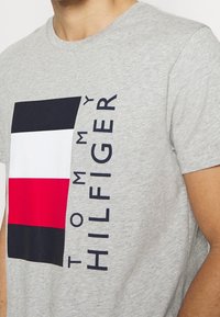 Camiseta de algodón gris con un diseño gráfico audaz que presenta secciones en azul marino, blanco y rojo, y "TOMMY HILFIGER" impreso en texto azul marino.