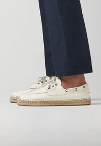 Zapatos de cuero blanco con suela de yute, que presentan ojales plateados y cordones grises, sobre pantalones oscuros, mostrando un diseño casual.