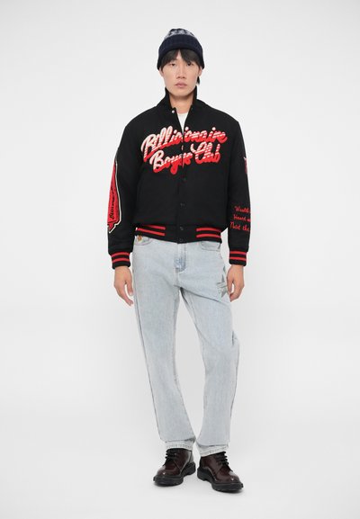 Casaco varsity preto com letras vermelhas, jeans de ganga cinza e botas castanhas com atacadores. O casaco possui bordados e detalhes risca.