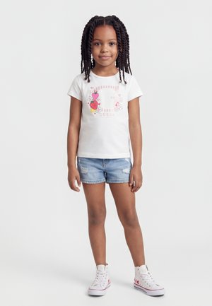 Jeune fille aux cheveux tressés portant un t-shirt blanc à motif floral, un short en denim et des baskets montantes blanches sur un fond uni.