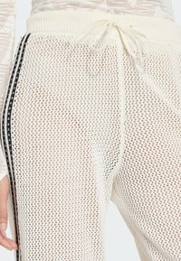 Biele sieťovinové nohavice s nastaviteľným pásom na šnúrky, čiernymi pásmi po stranách a voľným strihom. Sieťovina má vzor diamantov a jemnú textúru.