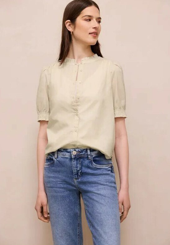 Femme portant un chemisier crème à manches courtes avec des détails à boutons et un jean bleu taille haute, debout devant un fond beige uni.