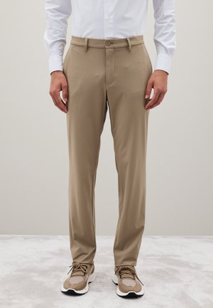 Pantalones de vestir beige hechos de un tejido ligero con un corte recto, que cuentan con cierre de botón y trabillas para cinturón, combinados con zapatillas beige.