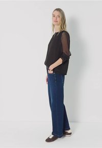 Haut noir transparent à manches longues, associé à un jean bleu à jambes larges et des baskets marron avec des lacets blancs. Fond blanc.