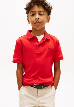 Berniukas su garbanotais plaukais, dėvintis raudoną polo marškinėlį, baltą marškinėlį po apačia, smėlio spalvos šortus ir juodą diržą, stovintis su rankomis kišenėse.