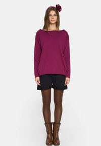 Bordowa bluzka z długim rękawem i szerokim dekoltem, w połączeniu z czarnymi szortami. Brązowe botki do kostki oraz pasująca opaska na głowę z akcentem w postaci kwiatu.