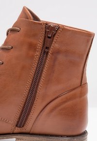 Bottine en cuir marron avec fermeture éclair latérale, lacets couleur tan, et petit talon empilé, présentée sur un fond blanc.