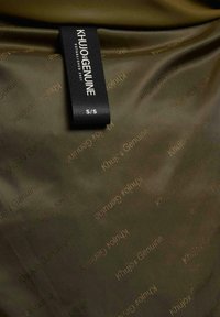 Oliven grøn stof med gentaget "Khujo Genuine" tekstmønster og et sort tøjlabel med teksten "KHUJO GENUINE ESTABLISHED 2021 S/S."