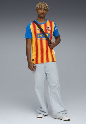 Camiseta de fútbol del Valencia CF con rayas verticales rojas y amarillas, mangas azules, logo en el pecho, combinada con pantalones de mezclilla azul claro y zapatillas blancas.