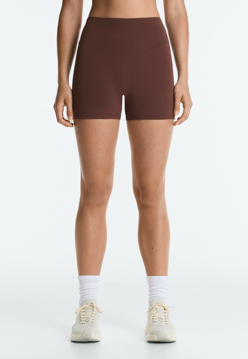 Femme portant un short de sport marron taille haute, des chaussettes blanches et des chaussures de course beiges, debout sur fond blanc.