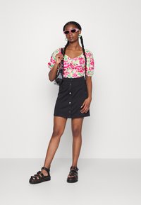 Top estampado floral com mangas bufantes em rosa e verde, combinado com uma saia preta de botão à frente e sandálias chunky pretas. Os acessórios incluem óculos de sol.