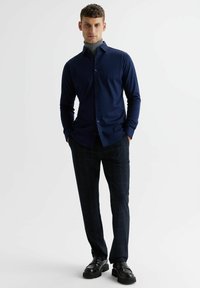 Granatowa koszula z długim rękawem z zapięciem na guziki, zestawiona z kraciastymi spodniami w ciemnoniebieskie i zielone odcienie. Czernie obuwie dopełniają stylizację.