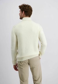 Crème-kleurige gebreide trui met hoge kraag en geribbelde zoom, gecombineerd met beige broek. Zachte textuur en aansluitend ontwerp.