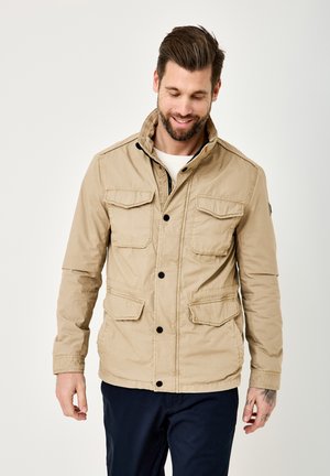 Smiling man with beard wearing beige jacket with four flap pockets and black buttons, standing against light background looking down.