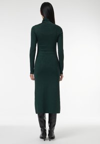 Robe verte ajustée à manches longues avec un col montant. Fabriquée en tissu texturé et scintillant. Portée avec des bottes noires jusqu'au genou.