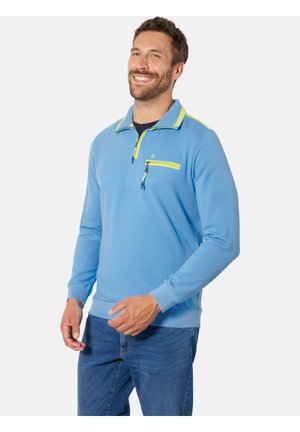 Mann lächelt, trägt einen hellblauen Viertelreißverschluss-Pullover mit neon-gelber Umrandung und Brusttasche, kombiniert mit blauer Jeans, steht aufrecht.
