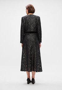 Juodas nėrinių suknelė su ilgu, laisvai krentančiu sijonu, papuoštu sudėtingais sidabriniais gėlių raštais, ilgomis rankovėmis ir prigludusia juosmens dalimi. Baigta juodais aukštakulniais.