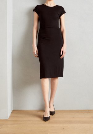 Woman wearing a fitted black midi dress with cap sleeves, black pointed-toe heels, a pearl necklace, and silver rings, standing on a wood floor.
