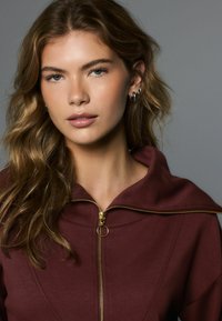 Bordo spalvos gobtuvu užsegamas švarkas su auksiniu užtrauktuku ir apskritu traukimo žiedu; minkštos medžiagos ir laisvo silueto.