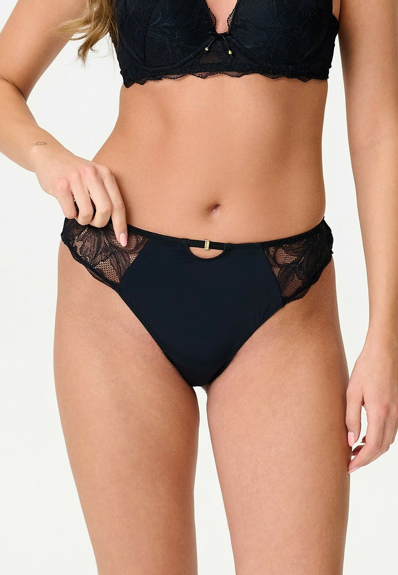 Femme portant un soutien-gorge en dentelle noire et une culotte en dentelle noire assortie, tenant le côté de la culotte de la main gauche.