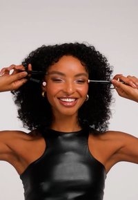 To svarte sminkepenner holdt av en person med krøllete hår som smiler og har på seg en svart ermeløs topp. Bakgrunnen er ensfarget hvit.