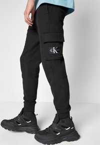 Svarta cargojoggers med ribbad detaljering, inklusive sidofickor och en patchlogotyp. Kombineras med svarta texturerade vandringsskor.