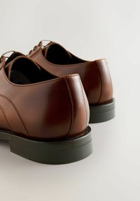 Brązowe skórzane buty z błyszczącym wykończeniem, okrągłym obcasem i teksturowanymi szarymi podeszwami. Cechuje je staranne szycie i gładkie linie.