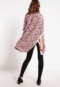 Mørkerød poncho med geometriske mønstre, med åbent front og cremefarvede accenter, båret over sorte leggings og ankelstøvler.