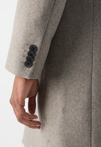 Manga de abrigo de lana gris que muestra cuatro botones negros, tejido texturizado y un ajuste slim. Mano descansando en la costura lateral del abrigo.