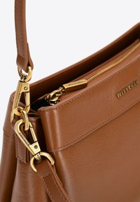 Brązowa skórzana torebka o gładkiej fakturze, z złotymi detalami, zamkiem błyskawicznym i strukturalnym kształtem. Logo widoczne z boku.