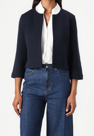 Femme portant une veste courte bleu marine sur une chemise blanche boutonnée et un jean bleu taille haute, debout devant un fond uni.