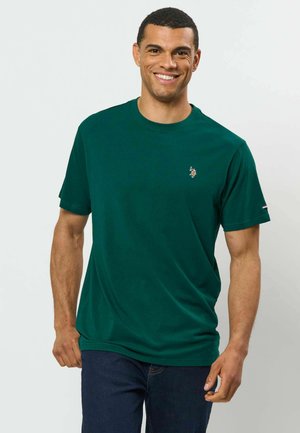 Homme souriant portant un t-shirt à manches courtes vert foncé avec un petit logo brodé sur la poitrine et un jean bleu foncé.