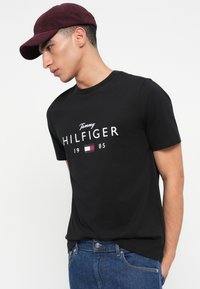 Mees, kes kannab marooni mütsi, musta Tommy Hilfiger'i t-särki logoga ja siniseid teksaseid, seisab valge tausta ees, vaadates vasakule.
