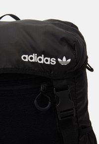 Černý batoh s měkkým textilním povrchem, se bílým logem adidas a zipovou kapsou. Obsahuje nastavitelné popruhy a kovové detaily.