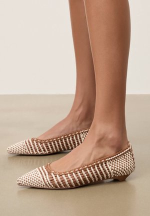 Brązowo-białe, tkane baleriny w szpic noszone na bose stopy, stojące na gładkiej beżowej podłodze z prostym jasnym tłem.