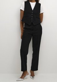 Gilet nero sartoriale con cinque bottoni, abbinato a pantaloni neri. Modella che indossa tacchi a punta argentati. Semplice T-shirt bianca sotto.