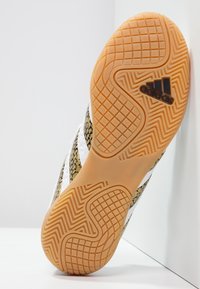 Calzado Adidas con un upper negro y dorado de textura y patrón, que presenta rayas blancas y una suela de goma con patrón de zigzag.