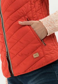 Czerwona pikowana kurtka z boczną kieszenią na zamek, teksturowanymi dzianinowymi rękawami oraz małą metką marki na dolnym brzegu.