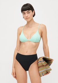 Mintskozeleni rebrasti bikini top s podesivim trakama uparen s crnim rebrastim bikini gaćicama visoki struk. Drži se višebojni tkaninasti ručnik.