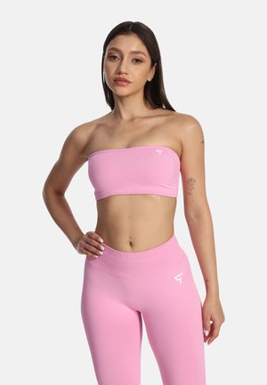 Kvinde iført matchende pink bandeau top og højtaljede leggings, stående med den ene hånd på hoften mod hvid baggrund.