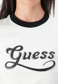 Hvit t-skjorte med svart ribbekrage som har en stor svart stilisert "Guess"-logo over brystet. Mykt bomullsstoff.