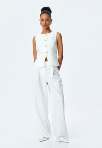 Weißes tailliertes Weste mit goldenen Knöpfen, kombiniert mit lockeren weißen Hosen. Glatter Stofftextur, minimalistisches Design, schlicht und dennoch elegant.